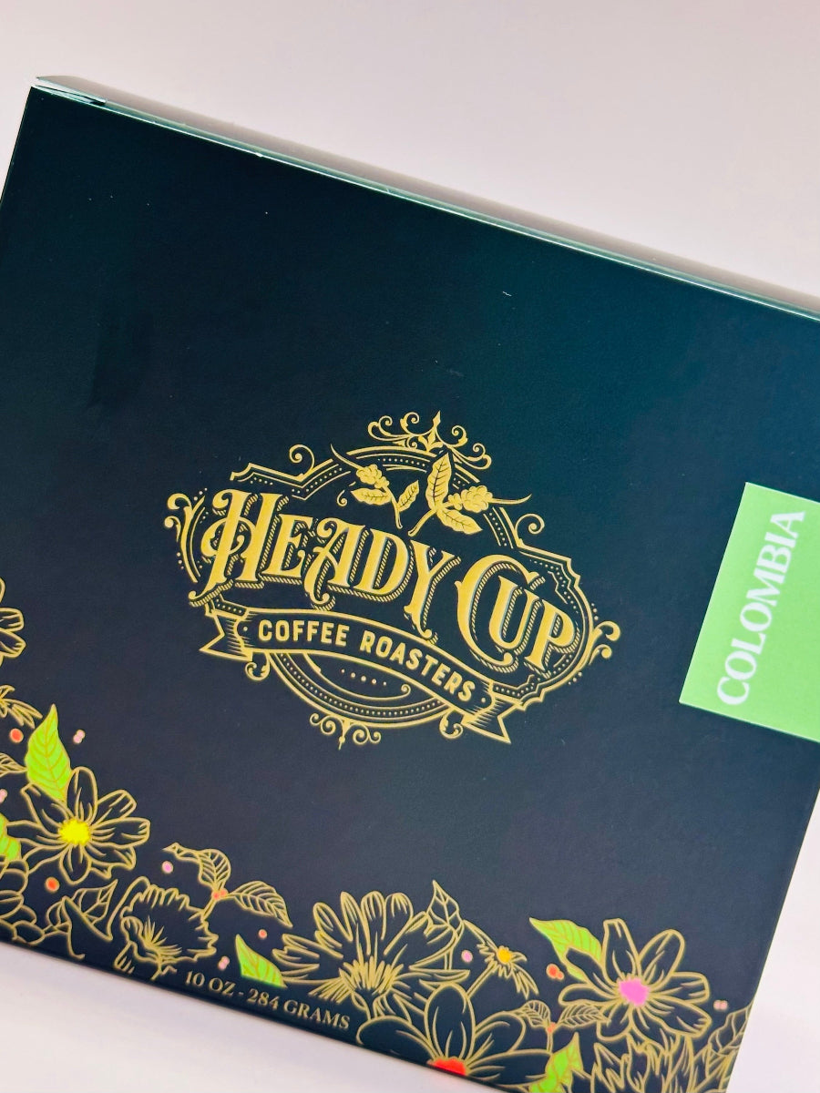 A close-up of a coffee box from Heady Cup Coffee Roasters, featuring a dark black background with ornate gold foil lettering and floral illustrations along the bottom. A bright green label on the right side reads 'COLOMBIA'.