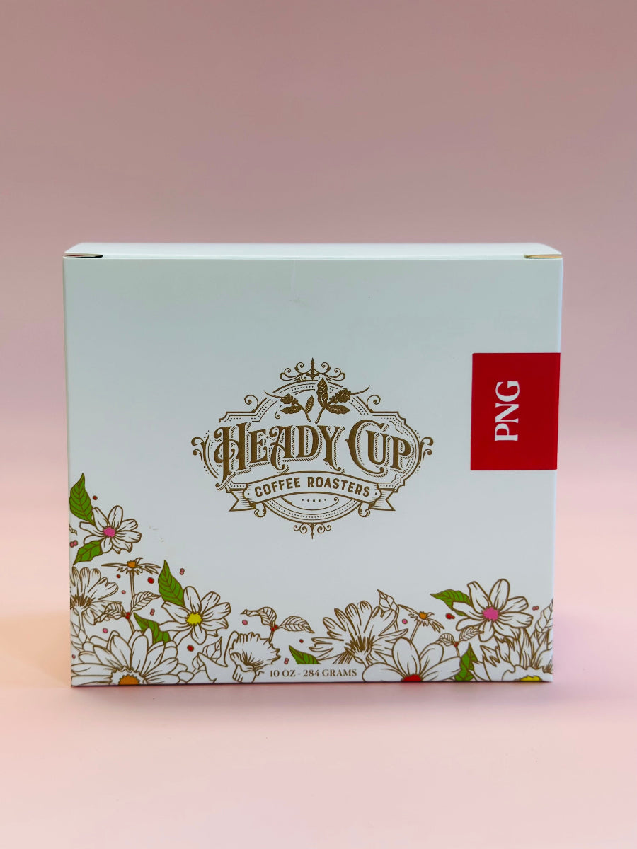 A single-origin coffee box from Heady Cup Coffee Roasters, decorated with gold floral illustrations and a red vertical label reading 'PNG' on the right side, set against a soft pink gradient background.