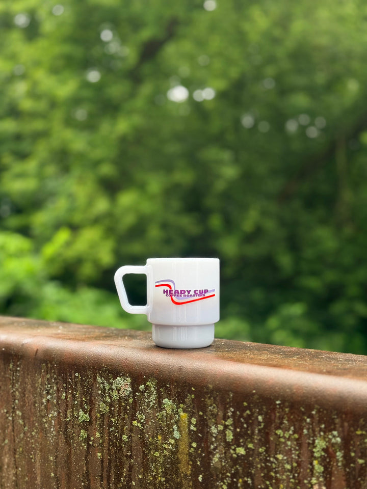A mist blue CreatedCo. Milk Glass with a red and blue Heady Cup Coffee Roasters logo on the front. The glass is pictured on the railing of a bridge with trees and a stream in the background. 