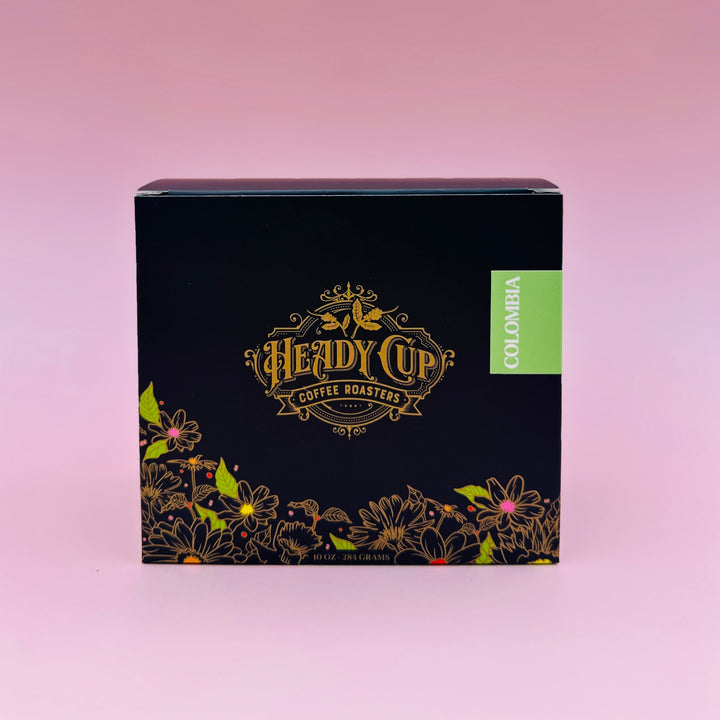 A square black coffee box for Heady Cup Coffee Roasters, featuring gold foil lettering and a green 'Colombia' label, set against a solid pink background.