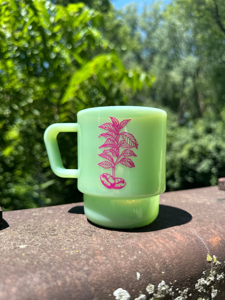 A jade green CreatedCo. Milk Glass with a pink Heady Cup Coffee Roasters tree on the front. The glass is pictured on the railing of a bridge with trees and a stream in the background. 