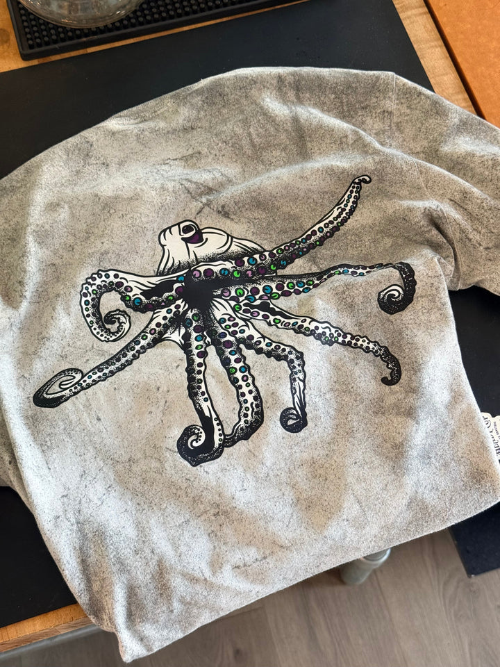 A Comfort Colors color blast t-shirt featuring a large black Heady Cup Coffee Roasters Flo octopus graphic with green and blue gem-like accents on its tentacles, laid flat on a dark surface.