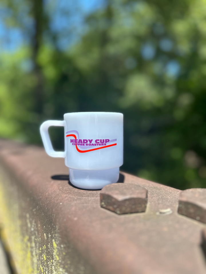A mist blue CreatedCo. Milk Glass with a red and purple Heady Cup Coffee logo Roasters on the front. The glass is pictured on the railing of a bridge with trees and a stream in the background. 