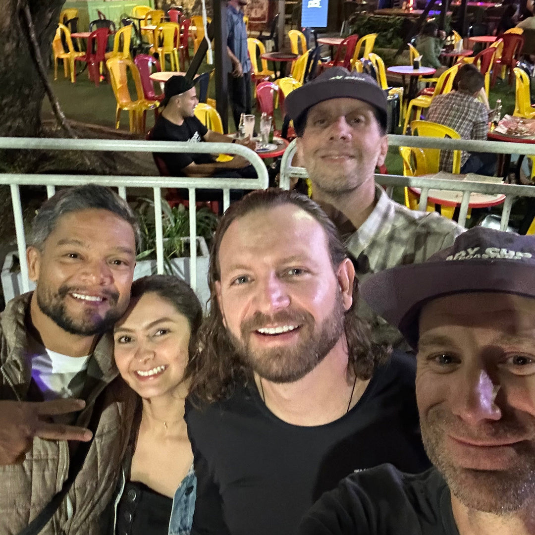 A group of five friends taking a selfie at an outdoor night market or restaurant. The background features colorful yellow and red chairs, tables, and warm ambient lighting.