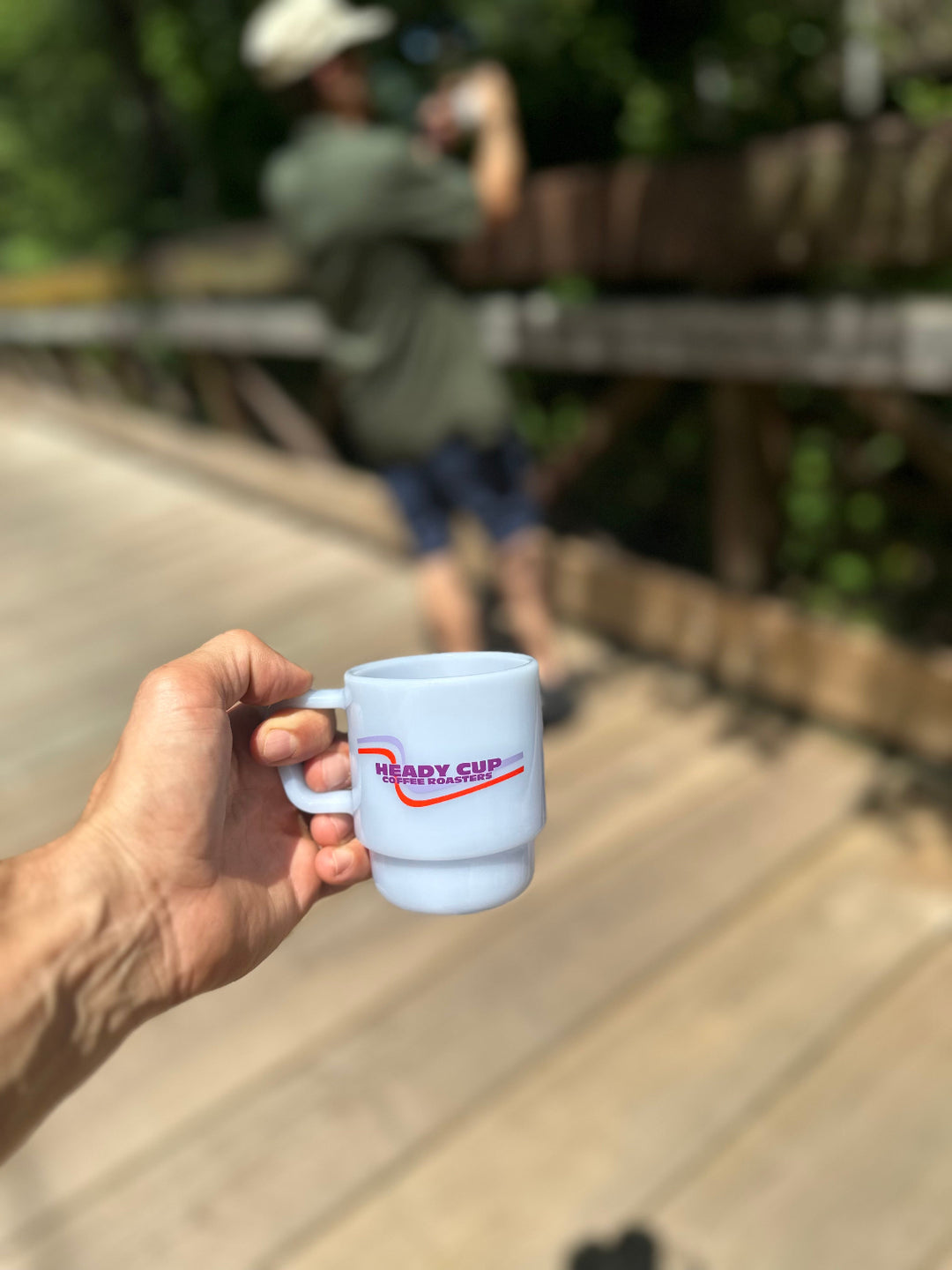 A mist blue CreatedCo. Milk Glass with a red and purple Heady Cup Coffee Roasters logo on the front. The glass is pictured on a bridge with trees and a man in the background. 