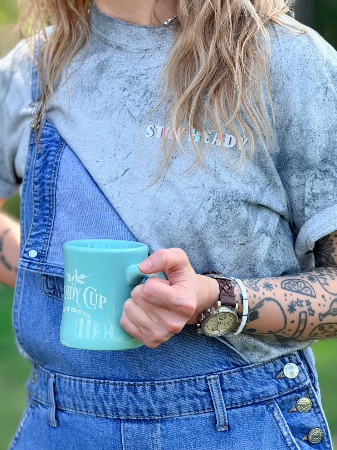 Grace wearing a gray Comfort Colors color blast t-shirt with 'STAY HEADY' printed on the chest and blue denim overalls, holding a mint green ceramic mug that says 'Heady Cup Coffee Roasters." 