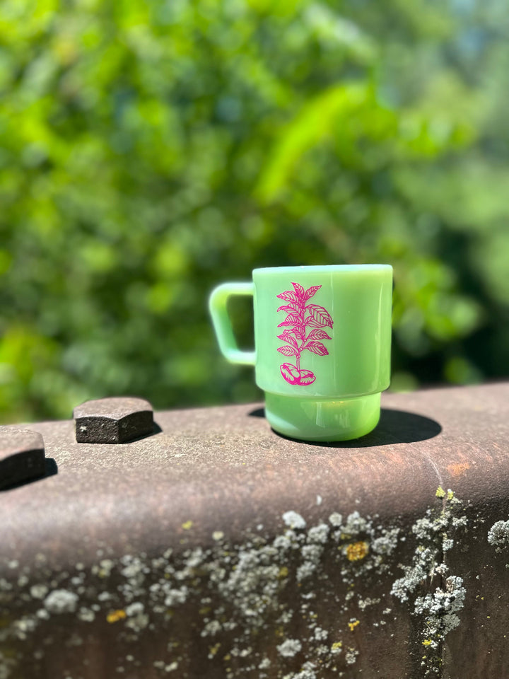 A jade green CreatedCo. Milk Glass with a pink Heady Cup Coffee Roasters tree on the front. The glass is pictured on the railing of a bridge with trees and a stream in the background. 