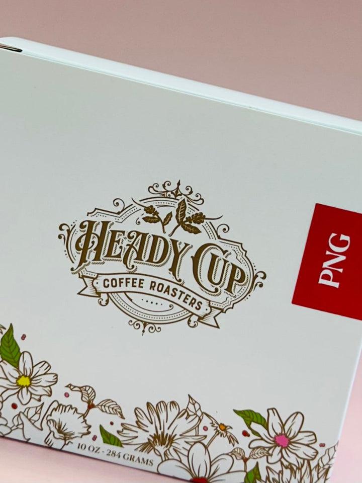A close-up of a white coffee roaster box featuring a decorative gold 'Heady Cup Coffee Roasters' logo, floral line art along the bottom, and a red square label with 'PNG' on the right side.