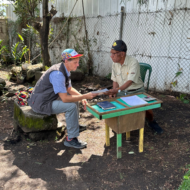 Two people sitting at a small wooden table outdoors with a fence and trees in the background.