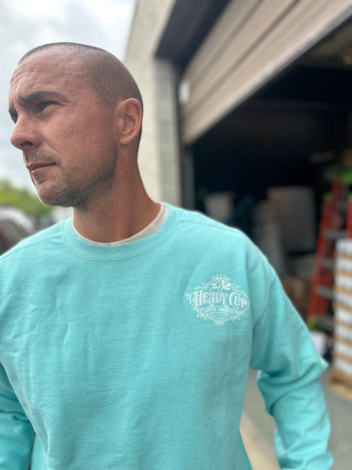 Man wearing a light blue Heady Cup Coffee Roasters long-sleeve shirt with a logo, standing outdoors.