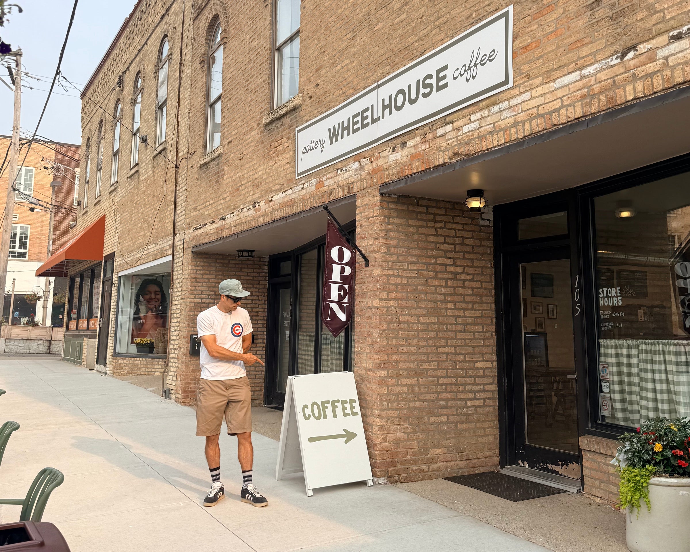Michael Kivland stands on the sidewalk in front of Wheelhouse Coffee on a brick street building, holding a phone. The storefront has a white sign reading 'WHEELHOUSE coffee' and a white A-frame sandwich board that says 'COFFEE' with an arrow.