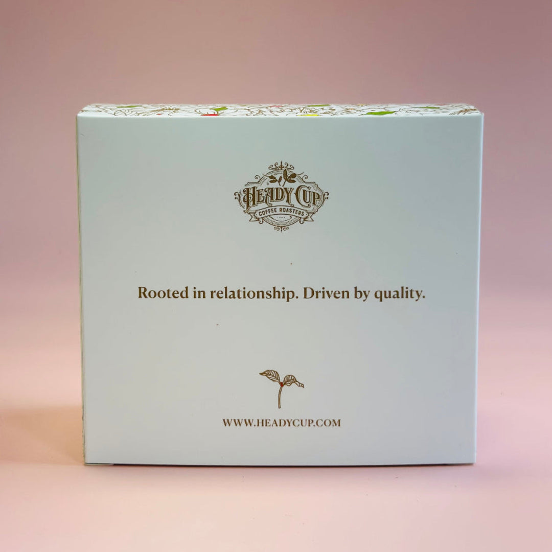 A square product box for 'The Herdy Cup' by Heady Cup Cakes, featuring a pale blue front with gold text and logo, set against a soft pink background.