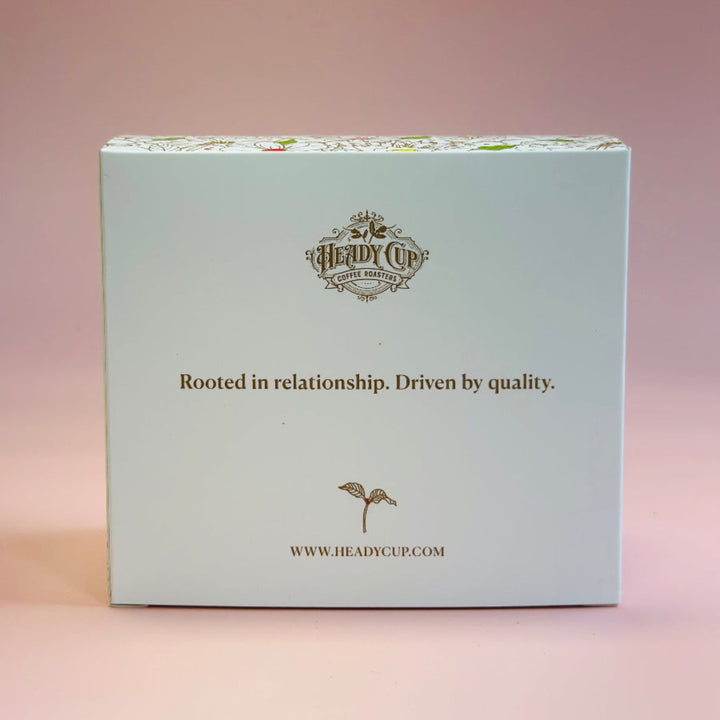 A square product box for 'The Herdy Cup' by Heady Cup Cakes, featuring a pale blue front with gold text and logo, set against a soft pink background.