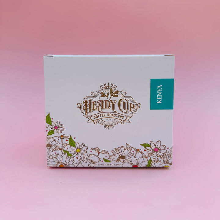 A white rectangular box for Heady Cup Coffee Roasters, featuring a gold logo, a teal 'KENYA' label, and a floral border at the bottom, set against a soft pink background.