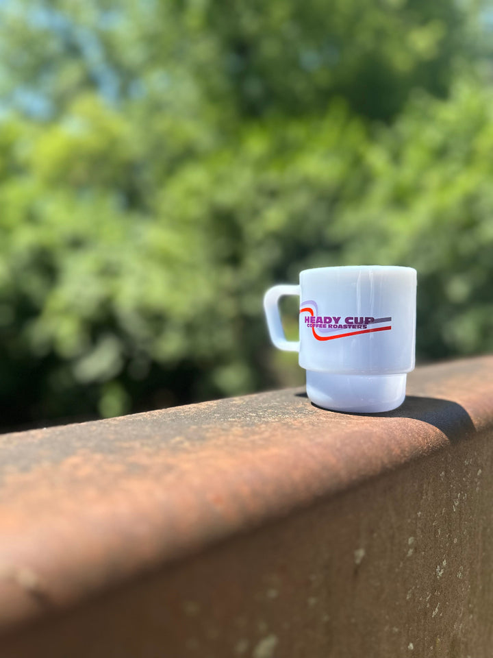 A mist blue CreatedCo. Milk Glass with a red and purple Heady Cup Coffee Roasters on the front. The glass is pictured on the railing of a bridge with trees and a stream in the background. 