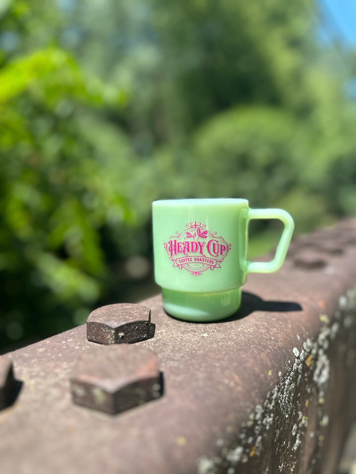 A jade green CreatedCo. Milk Glass with a pink Heady Cup Coffee Roasters logo on the front. The glass is pictured on the railing of a bridge with trees and a stream in the background. 