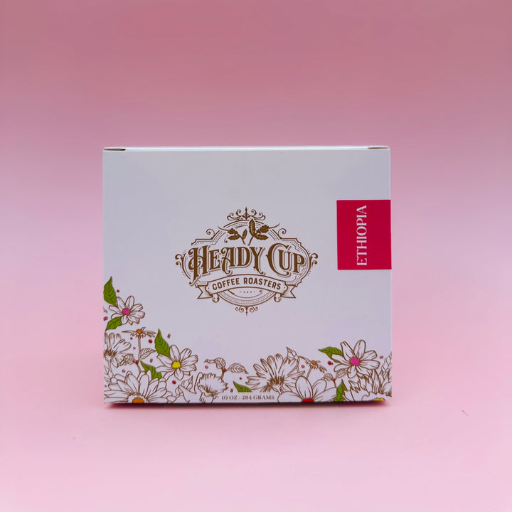 A white single-origin box for Heady Cup Coffee Roasters from Ethiopia, featuring gold foil branding, white floral illustrations, and a red vertical label, set against a solid pink background.