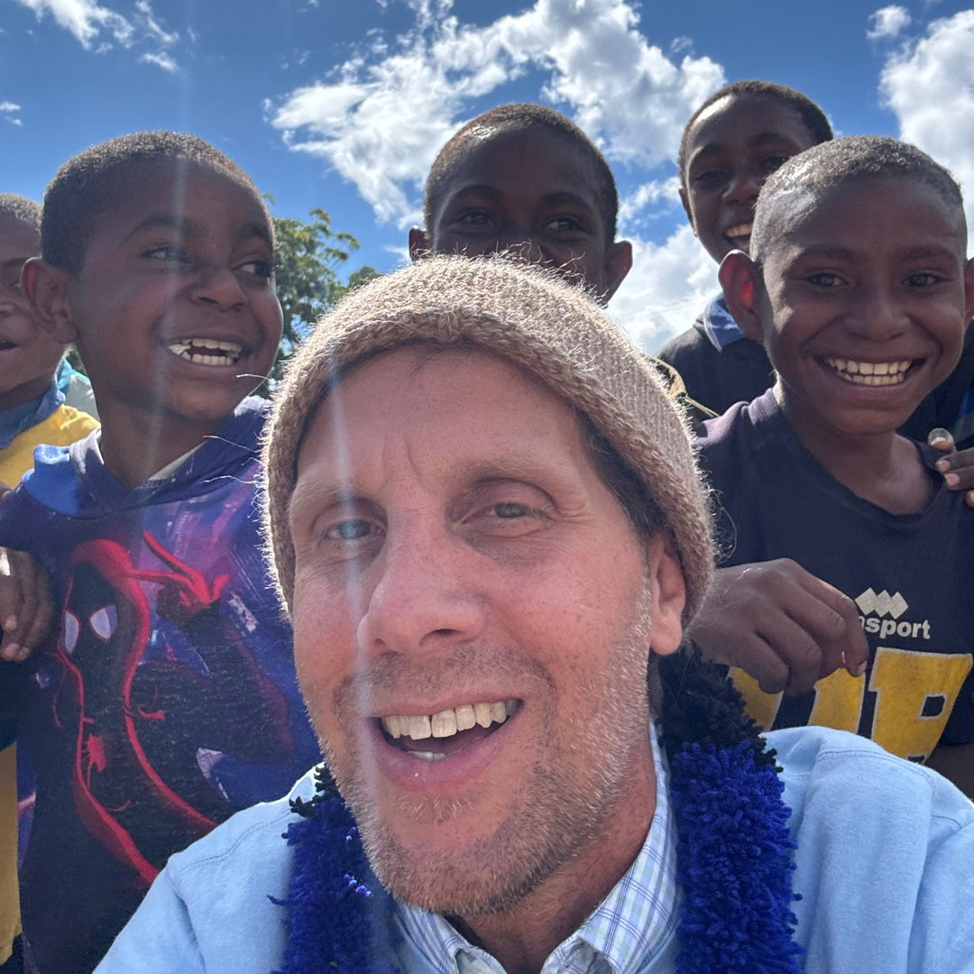 A smiling man in a beige knit beanie and blue flower lei stands outdoors with a group of young boys under a bright blue sky with white clouds. One boy on the right wears a dark shirt with yellow lettering.
