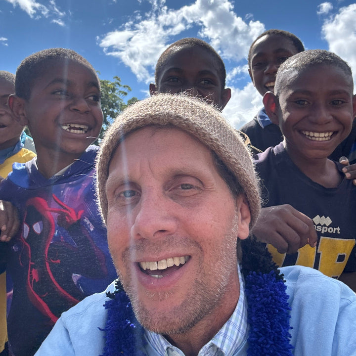 A smiling man in a beige knit beanie and blue flower lei stands outdoors with a group of young boys under a bright blue sky with white clouds. One boy on the right wears a dark shirt with yellow lettering.
