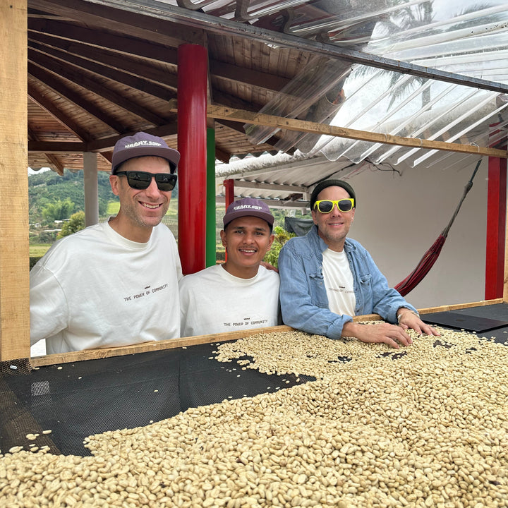 Three men, Michael Kivland, Marlon Bolaños, and Kurtis Kearby, wearing matching white UNBLND t-shirts and baseball caps stand behind a large spread of raw coffee beans under a rustic corrugated metal roof.