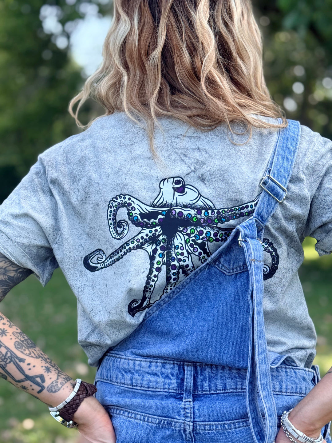 A woman viewed from behind wearing a gray Comfort Colors color blast t-shirt with a large black Heady Cup Coffee Roasters Flo octopus graphic on the back, styled with blue denim overalls outdoors.