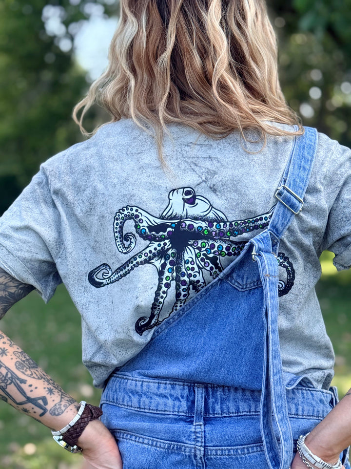 A woman viewed from behind wearing a gray Comfort Colors color blast t-shirt with a large black Heady Cup Coffee Roasters Flo octopus graphic on the back, styled with blue denim overalls outdoors.