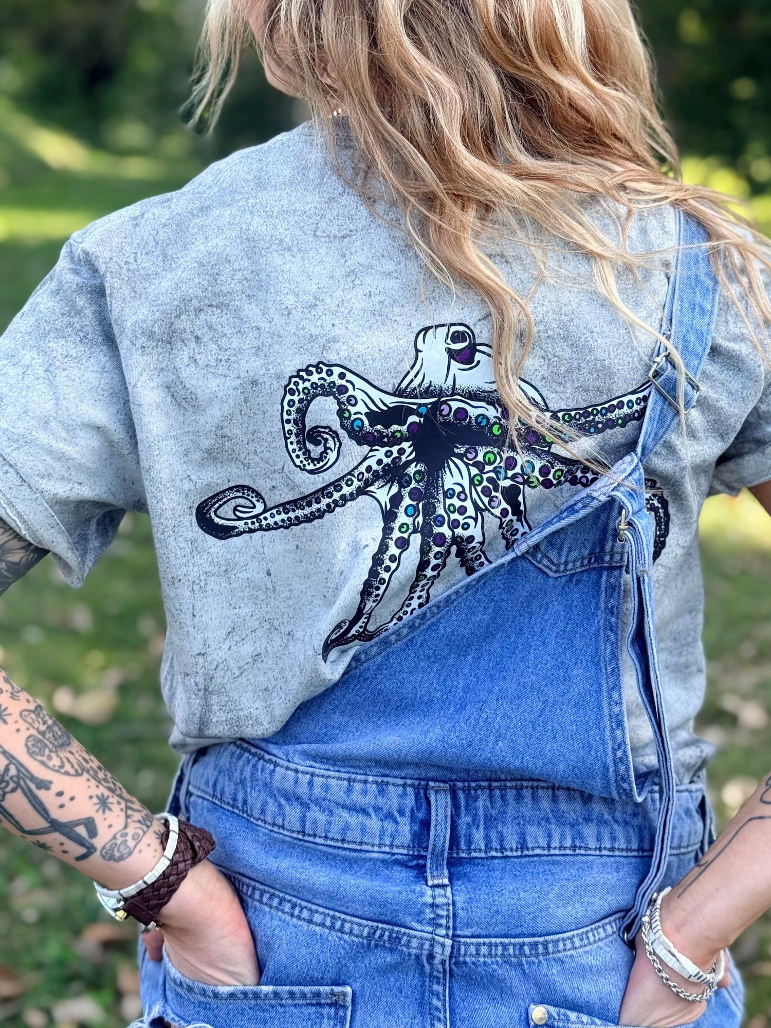 A woman viewed from behind wearing a gray Comfort Colors color blast t-shirt with a large black Heady Cup Coffee Roasters Flo octopus graphic on the back, styled with blue denim overalls outdoors.