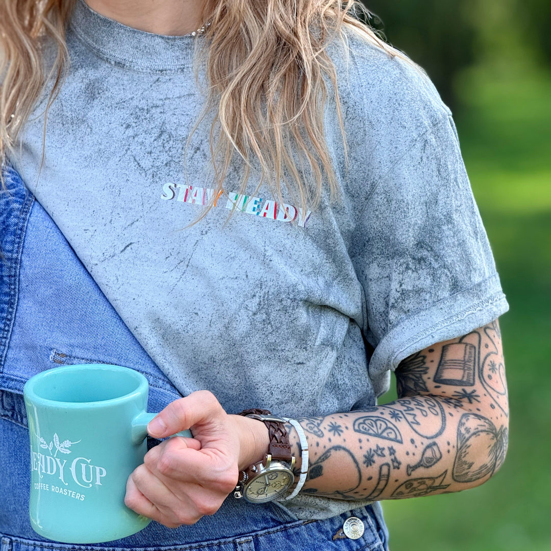 A close-up lifestyle shot of Grace wearing a Comfort Colors color blast t-shirt with 'STAY HEADY' printed on the chest, layered under blue denim overalls. The person is holding a light blue ceramic mug with 'Heady Cup Coffee Roasters' printed on it, set against a blurred green outdoor background.