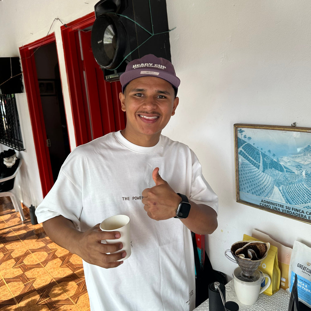 Marlon Bolaños wearing a white UNBLND t-shirt and a purple  Heady Cup Coffee Roasters cap stands in a home setting, holding a white coffee cup and giving a thumbs-up. A coffee maker and other kitchen items are visible on the counter behind him.