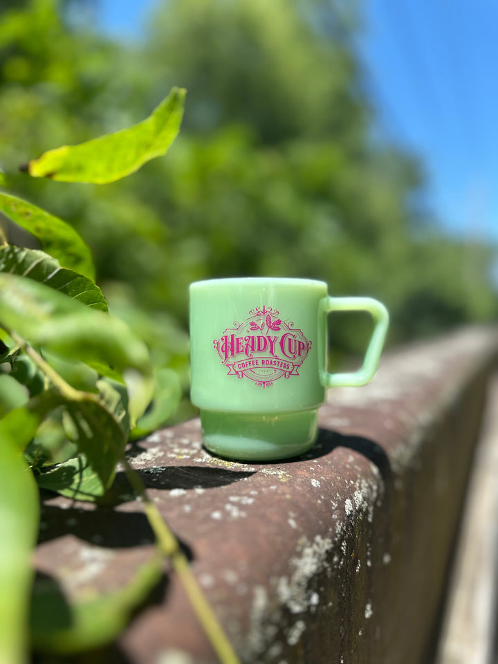 A jade green CreatedCo. Milk Glass with a pink Heady Cup Coffee Roasters logo on the front. The glass is pictured on the railing of a bridge with trees and a stream in the background. 