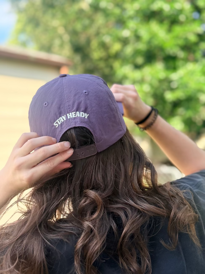 A woman wearing a purple Weld nylon hat with Heady Cup Coffee Roasters "Stay Heady" embroidered on the back of the hat. 