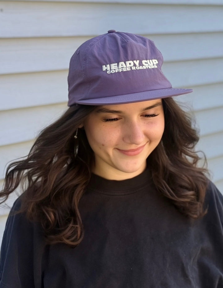 A woman wearing a purple Weld nylon hat with Heady Cup Coffee Roasters  embroidered on the front of the hat. 