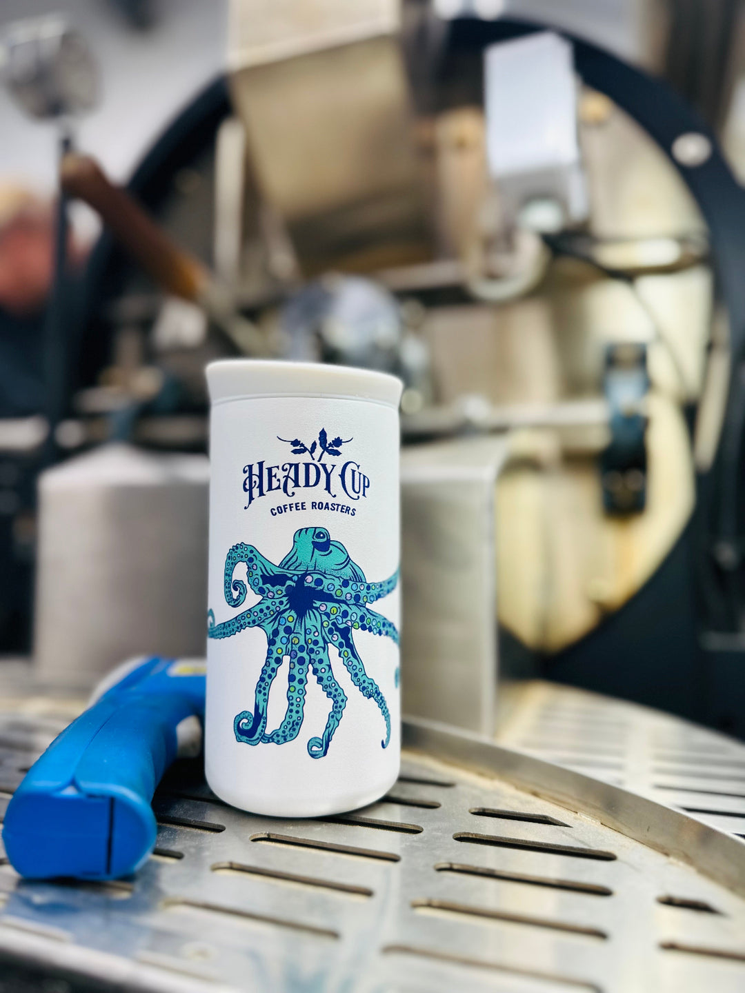 White CreatedCo NOMAD tumbler with blue octopus design and 'Heady Cup Coffee Roasters' branding in front of a coffee roaster