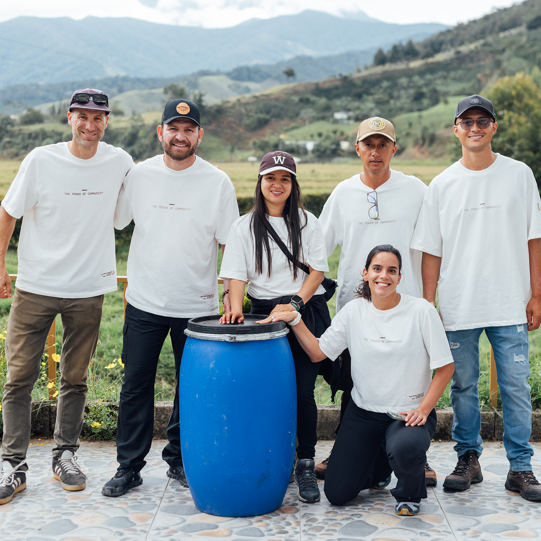 A team of coffee professionals standing behind a fermentation tank with coffee trees and mountains in the background. In the picture are Colombian coffee professionals, Sebastian Ramirez, Sofia Cuadros, and Yessica Parra. 