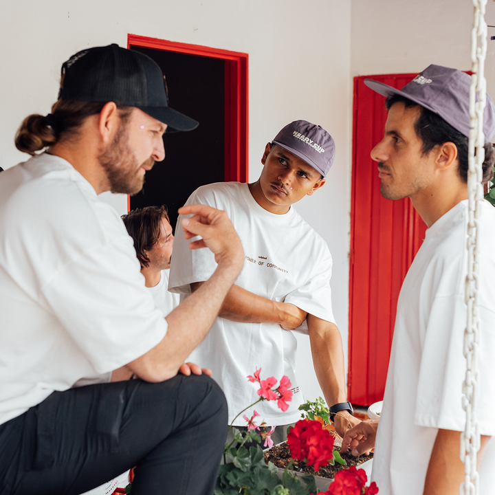 Three men in white t-shirts and caps converse in a room with red door frames and hanging white ropes, with flowers in the foreground.